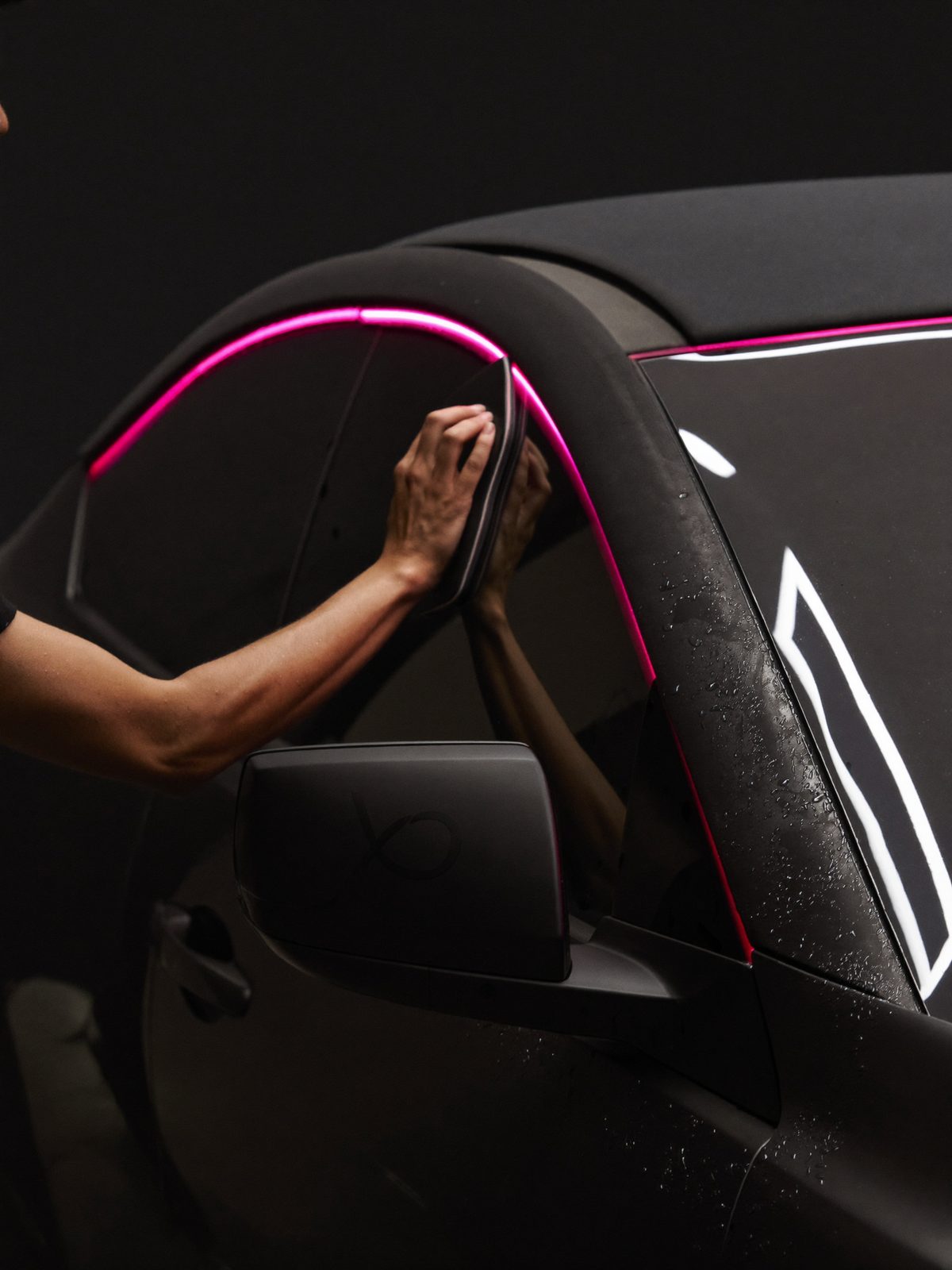 Installer smoothing window tint film onto car glass under pink shop light